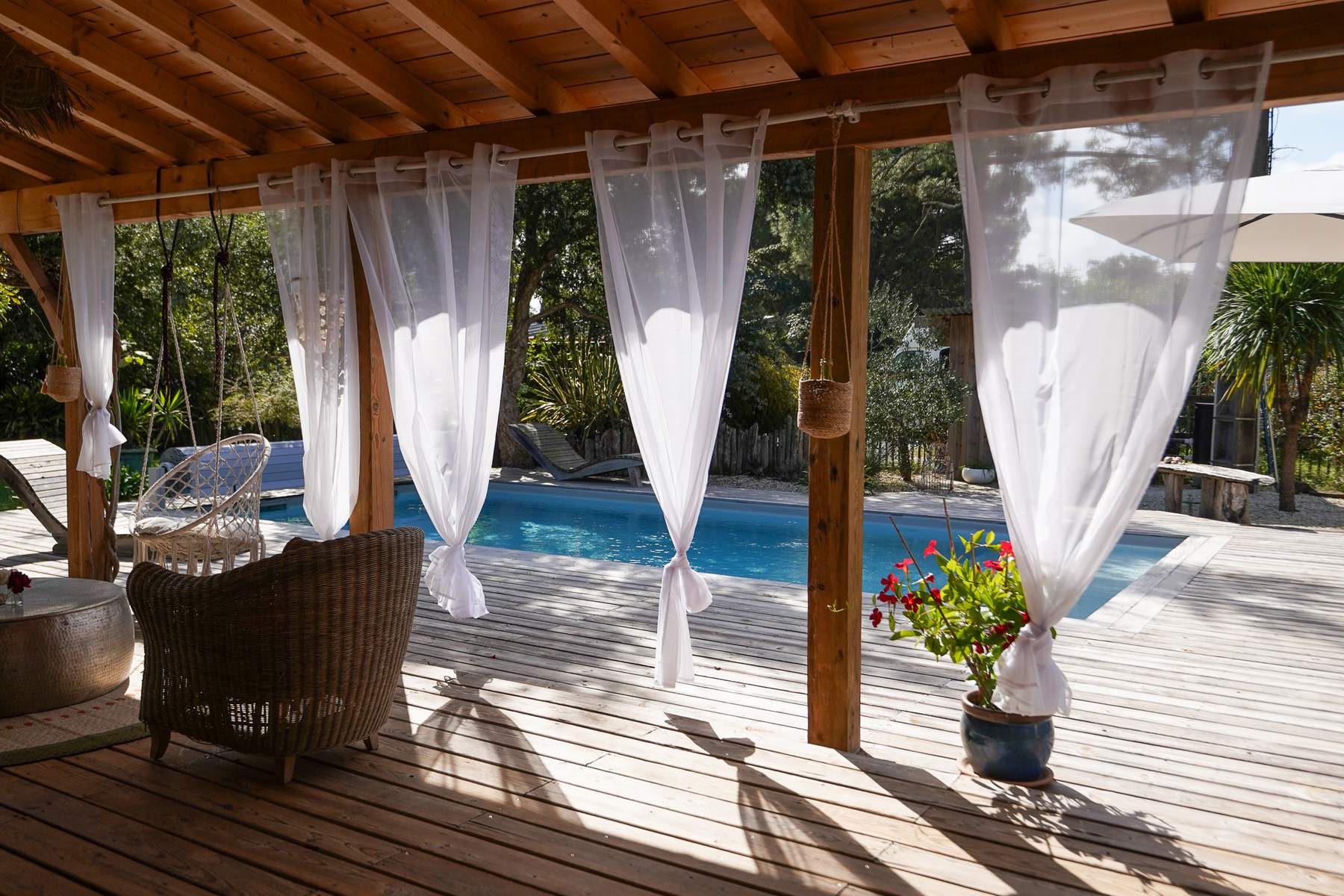 Rattansessel unter der Pergola mit weißen Vorhängen, mit Blick auf den beheizten Pool